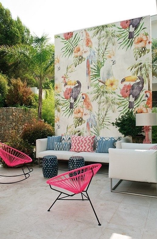 a colorful tropical patio with a white sofa and colorful pillows, hot pink chairs, a bright watercolor wall and little stools