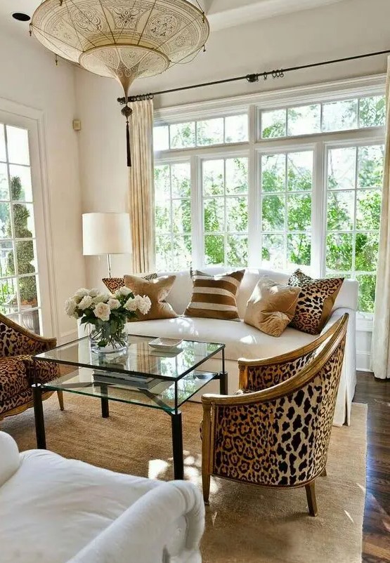 an exquisite living room done in neutrals, with a creamy sofa with printed pillows, leopard print chairs, layered rugs and a glass table