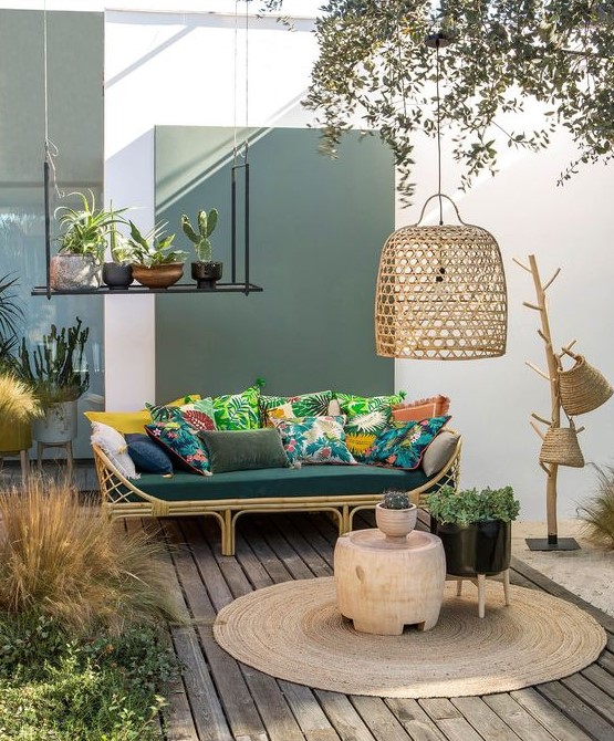 a welcoming tropical terrace with a rttan sofa, colorful pillows, a jute rug, wicker baskets and a wicker lampshade plus potted plants