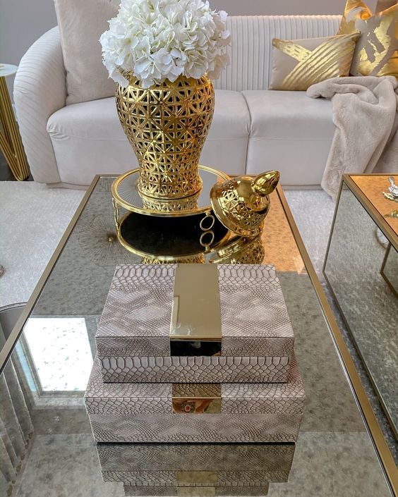 a glam space with a glass coffee table, pink snakeskin print boxes, a gold vase on a gold stand and white blooms