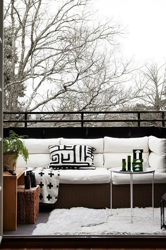a Nordic balcony with a wicker sofa with white upholstery, a wood and wicker side table, greenery and a glass side table
