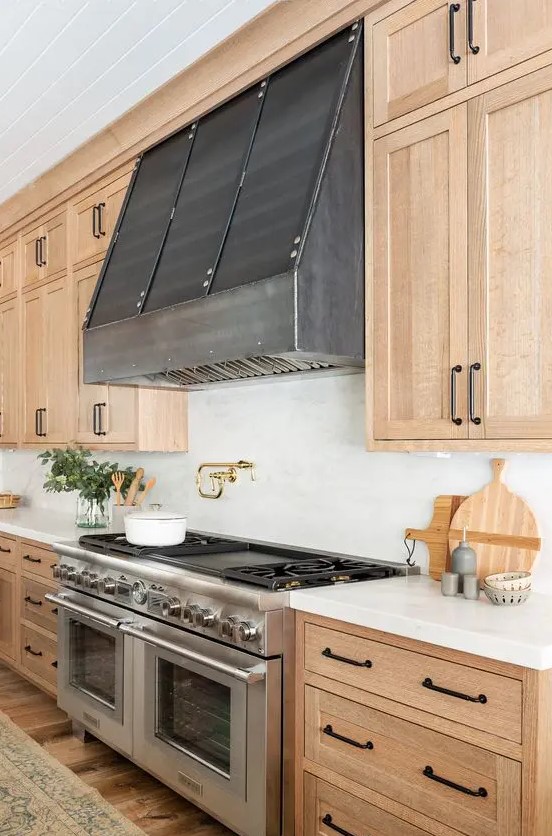 a chic light wood kitchen with shaker style cabinets, white stone countertops and a white stone backsplash, a metal hood and black handles