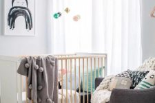 a cool Scandi nursery with a stained crib and a grey chair, muted and pastel color bedding, printed pillows and a pastel mobile