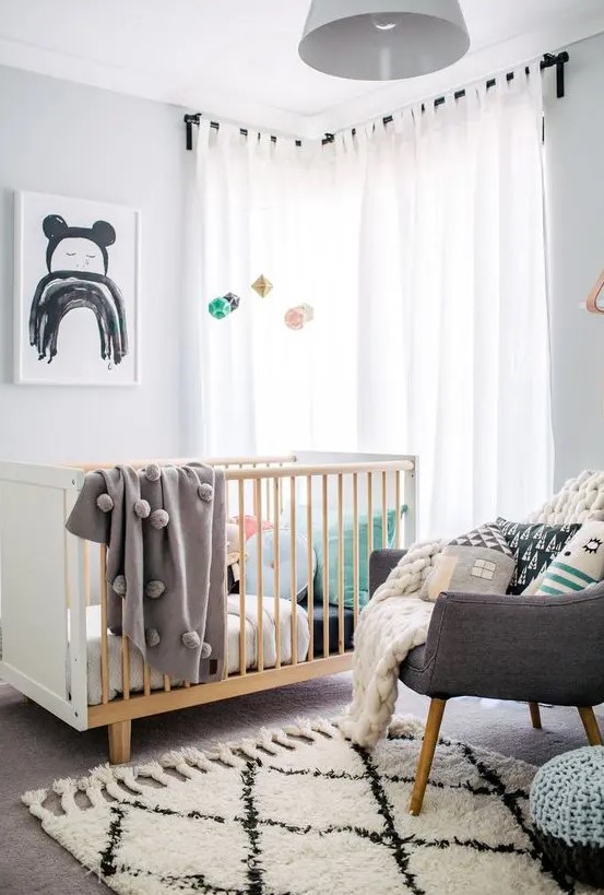 a cool Scandi nursery with a stained crib and a grey chair, muted and pastel color bedding, printed pillows and a pastel mobile