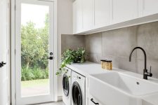 a laconic modern laundry with a concrete floor and walls, white shaker style cabinets, black handles and a door to the garden