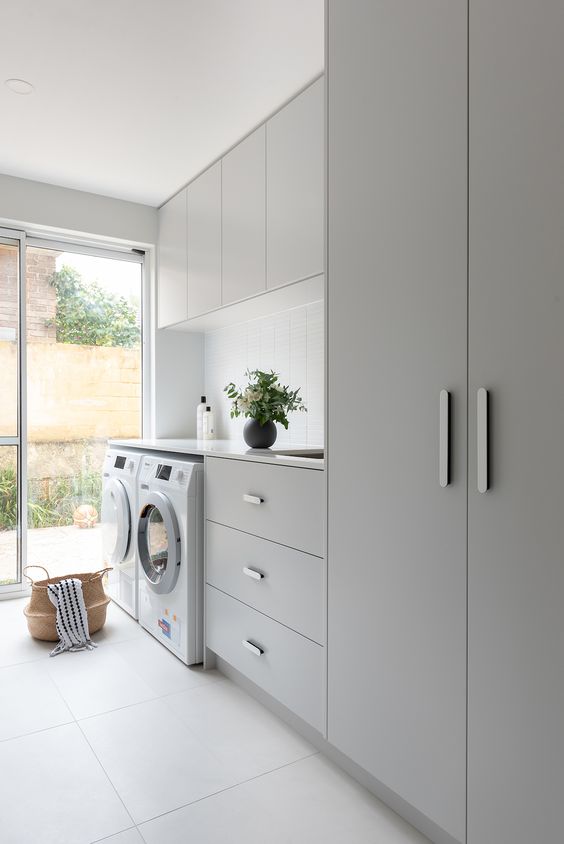 a lovely minimalist laundry with sleek grey cabinets and a wardrobe, a white skinny tile backsplash and neutral appliances is cool