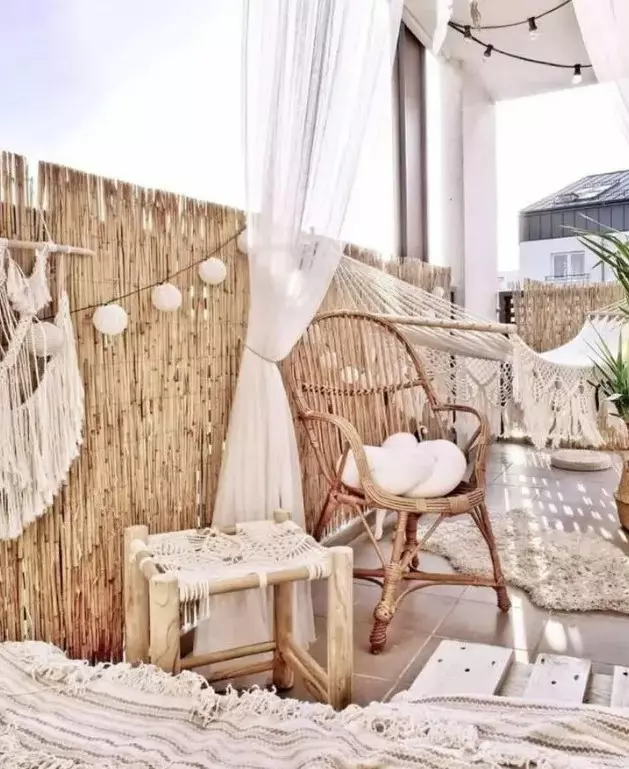 a lovely neutral boho balcony with rattan furniture, a hammock, a loveseat with a boho blanket, paper lamps, macrame and fringe is a very cozy space