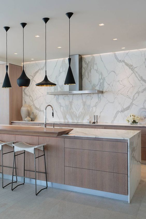 a minimalist kitchen with light-stained cabinets and a white marble wall and countertops, a cluster of black and gold pendant lamps