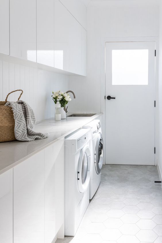 a minimalist white laundry with sleek and glossy cabinets, a stone countertop, a hexagon tile floor and brass fixtures