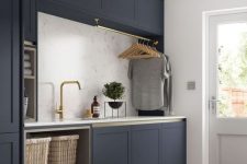 a modern farmhouse laundry with navy cabinets, a white stone countertop and a backsplash, gold fixtures and baskets for storage
