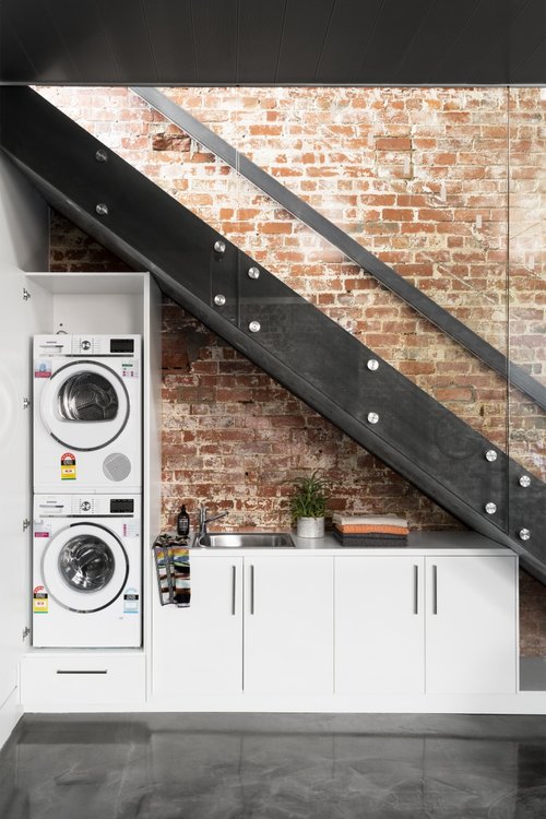 a modern industrial space with a brick wall and a darkened metal staircase, with a built-in washing machine and dryer, with storage cabinets