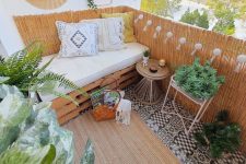 a neutral boho balcony with a crate mini sofa, lights and candles, potted greenery and pendant lamps, a tiled floor and a jute boho rug