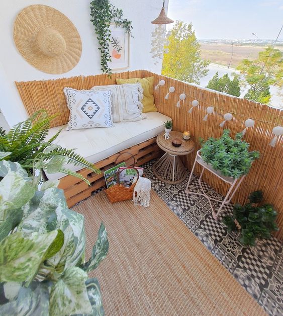 a neutral boho balcony with a crate mini sofa, lights and candles, potted greenery and pendant lamps, a tiled floor and a jute boho rug
