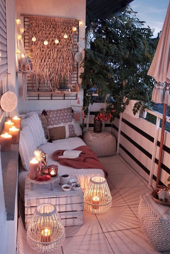 a neutral boho balcony with lights, candles, candle lanterns, a neutral sofa with lots of pillows, a crate with decor and greenery over the balcony