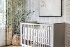 a relaxed Scandinavian nursery with printed wallpaper, a grey crib, an artwork, a mobile, a potted tree, layered rugs and striped curtains
