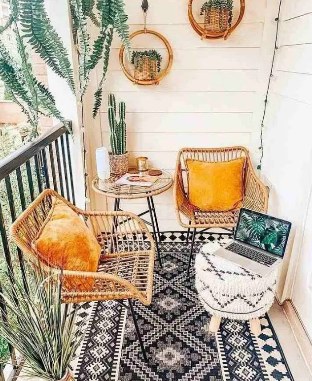 a small boho balcony with a black and white rug, rattan chairs with yellow pillows, a black and white pouf, potted plants and hanging planters