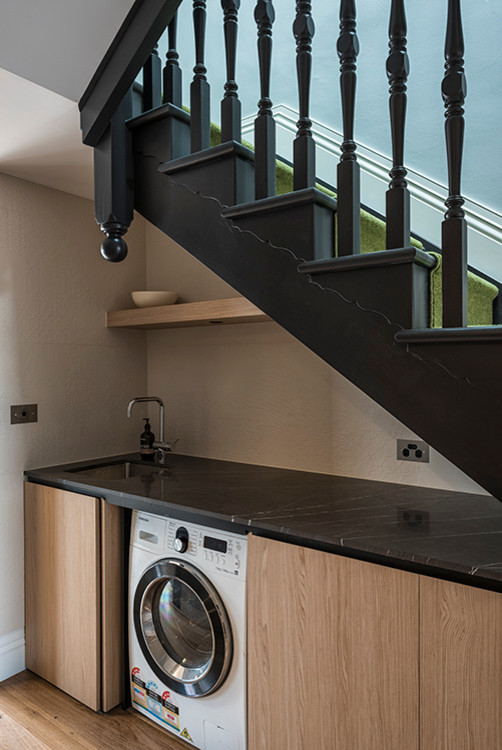 a stylish laundry under the stairts with a built-in black shelf, light-stained storage cabinets and a washing machine