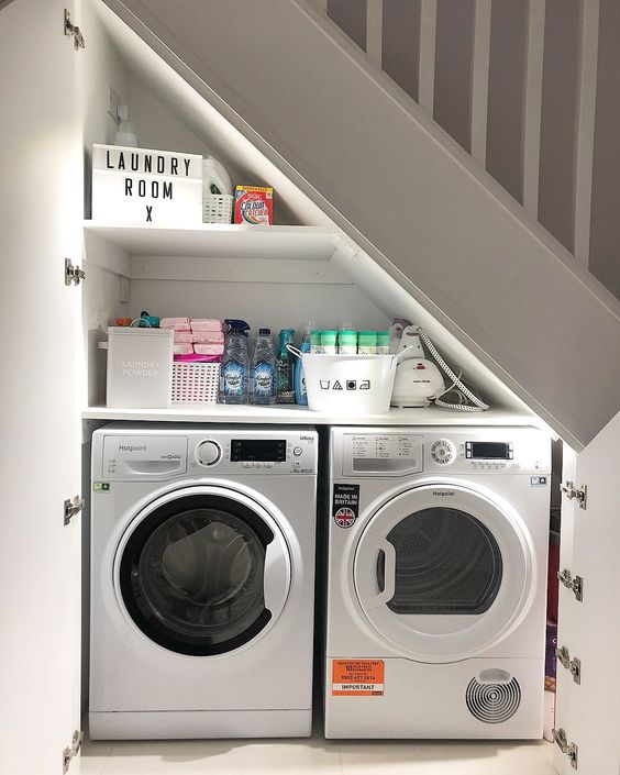 a tiny under the stairs laundry room with a built-in shelf and a washing machine and a dryer plus a couple of doors to hide it