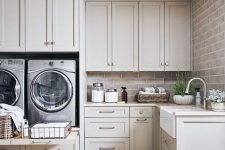 a welcoming grey farmhouse laundry with shaker style cabinets, taupe tiles on the walls, stainless steel appliances