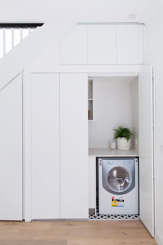 an under the stairs space with sleek white doors and a small laundry built-in, with a storage unit and a tiled floor is cool and functional