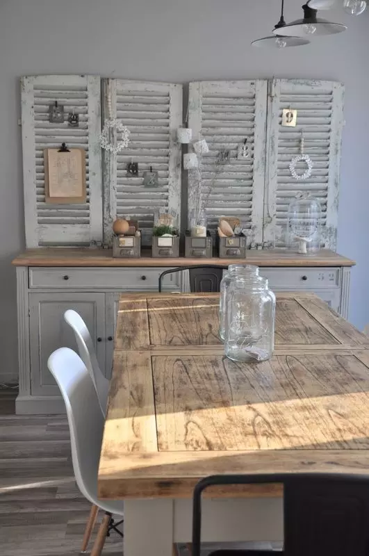 a farmhouse dining room with a table made of a door, white chairs, a grey sideboard, shabby chic shutters with various decor