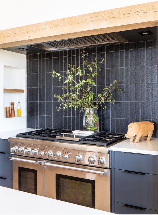 a grey kitchen with wooden beams, a black matte skinny tile backsplash and a metal cooker and hood