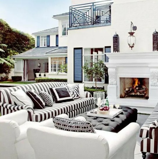 a lovely outdoor living room with a fireplace, striped black and white sofas, white chairs, a black upholstered ottoman and some greenery