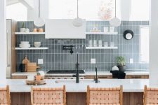a neutral stained wooden kitchen, white shelves and a hood, a blue skinny tile wall and pendant lamps