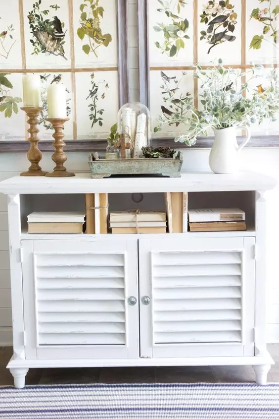 a cottage style white cabinet with shutter doors, candles in wooden candleholders, vintage books and greenery is ideal for a rustic space
