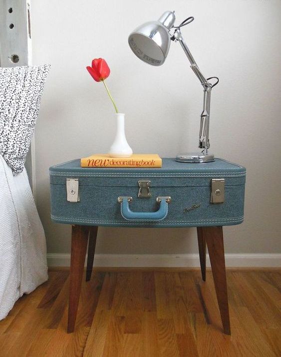 a stylish nightstand made of a blue fabric upholstered suitcase on legs, with a metal table lamp and a vase