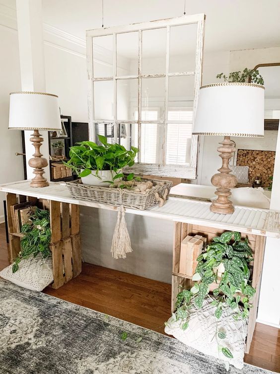 a lovely rustic console table made of crates and a shabby chic shutter, macrame pillows, greenery, books, table lamps and a basket with greenery
