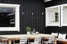 26 a Scandinavian terrace with a wooden deck, a stained corner bench, a stained wooden dining table and white metal chairs, lights over the table