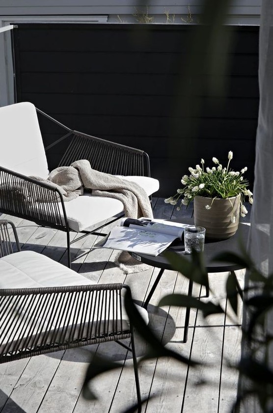 a small modern terrace with metal and wicker chairs, a black table, some simple blooms in a pot and a weathered deck