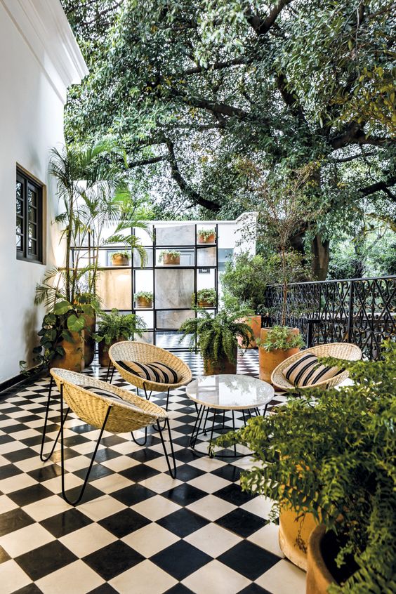 a welcoming terrace with black and white checked floor, a round table and woven chairs, potted greenery around and a storage unit