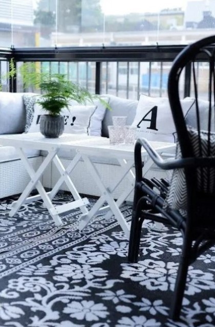 a black and white Scandinavian balcony with a printed rug, a white corner sofa, a black chair and white folding tables and greenery