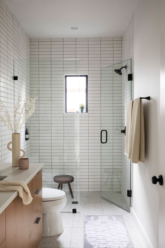 a delicate and welcoming bathroom clad with neutral skinny tiles, large terrazzo ones, black fixtures and a stained vanity plus a window for naturla light