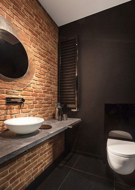 a moody and chic bathroom in black with a single red brick wall that makes the space stand out