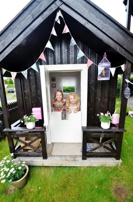 a Nordic black and white kids' playhouse with a pastel garland, a cage, potted blooms and a pink watering can is amazing for any kids' games
