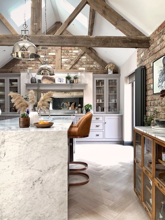 a barn kitchen with brick walls, grey shaker style cabinets, an elegant stone kitchen island with rust stools and aged wooden beams