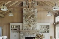 a barn living room with stained walls and a ceiling, with wooden beams, a fireplace clad with stone, neutral seating furniture and built-in storage units