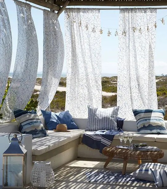 a beach patio with printed curtains, a bench with bright blue pillows, a wicker table and a candle lantern is very welcoming