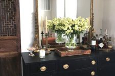 a beautiful black vintage dresser with gold knobs featuring a small home bar, a large mirror with a chic frame and some candleholders
