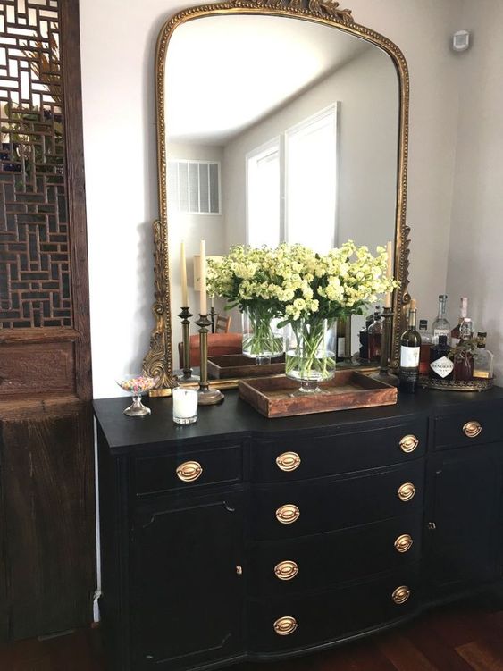 a beautiful black vintage dresser with gold knobs featuring a small home bar, a large mirror with a chic frame and some candleholders