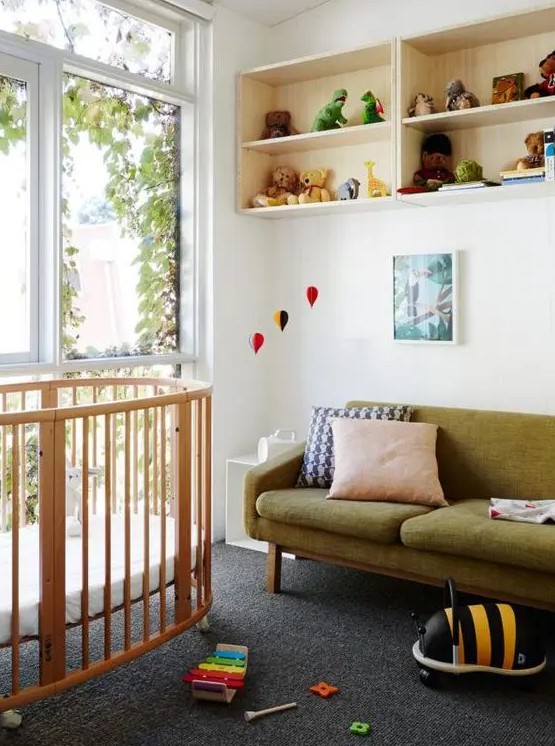 a beautiful mid-century modern nursery with open box shelves, a stained round crib, a green sofa, artworks, bright toys that double as decor
