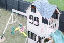 a black and white kids’ playhouse with a grey roof, a turquoise slide, swings, greenery growing and a checked rug is all cool