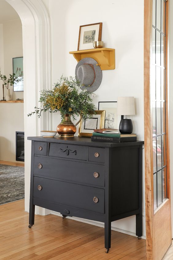 a black vintage dresser placed on casters, with artwork, greenery and a table lamp is placed in this awkward nook to make use of it