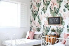 a boho desert shared girls’ bedroom with white metal beds, a jute rug, colored and printed pillows, a cactus print accent wall and a stained dresser