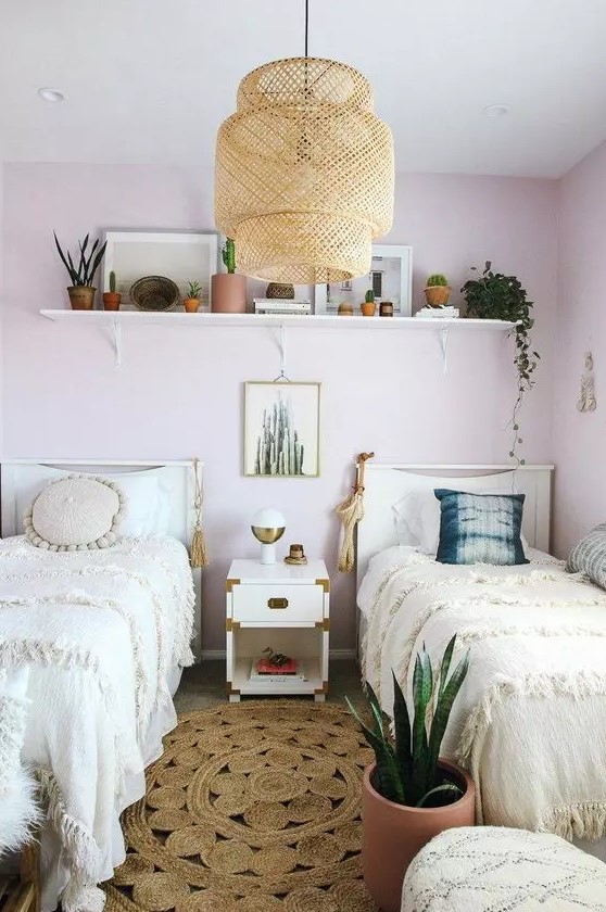 a boho shared girls' bedroom with light pink walls, white beds and a nightstand, a shelf with lots of plants, a woven pendant lamp