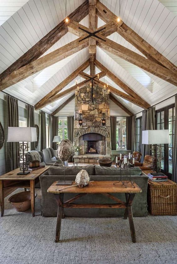a bold barn living room with wooden beams, a stone fireplace, graphite grey seating furniture and curtains, stained tables