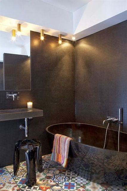 a bold bathroom with chocolate brown tiles, a black bathtub, a colorful tile floor, a floating sink and gilded lamps